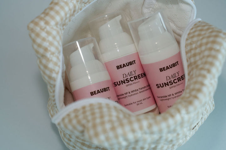 Three BEAUBIT Daily Sunscreen SPF 50 pump bottles with pink labels and white caps, arranged inside a beige and white gingham fabric pouch on a light blue-gray surface.