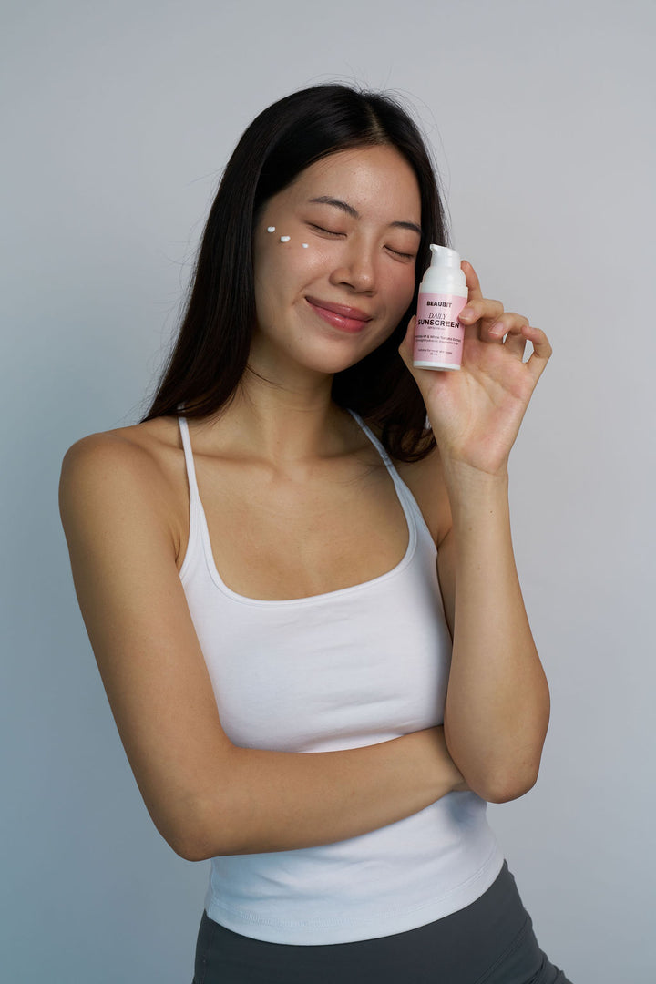 A woman with dark hair wearing a white tank top holds a small pink and white cosmetic bottle near her face, which has a few white dots on it, against a plain light gray background.
