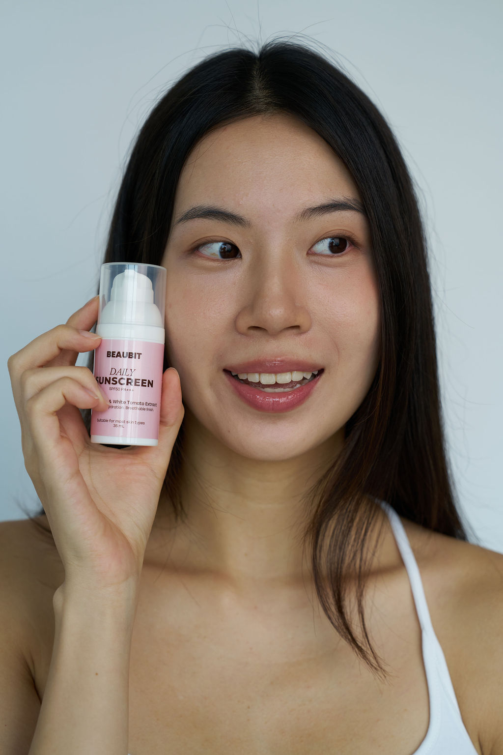 A woman holding a bottle of BUEABIT Sunscreen next to her face, wearing a white spaghetti strap top against a plain light gray background.