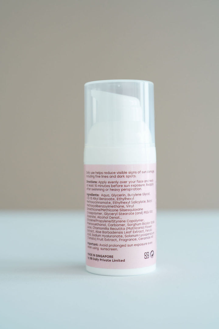 A white cylindrical cosmetic pump bottle with a clear cap, standing upright on a light gray surface against a neutral gray background. The bottle has a pale pink label with black text.