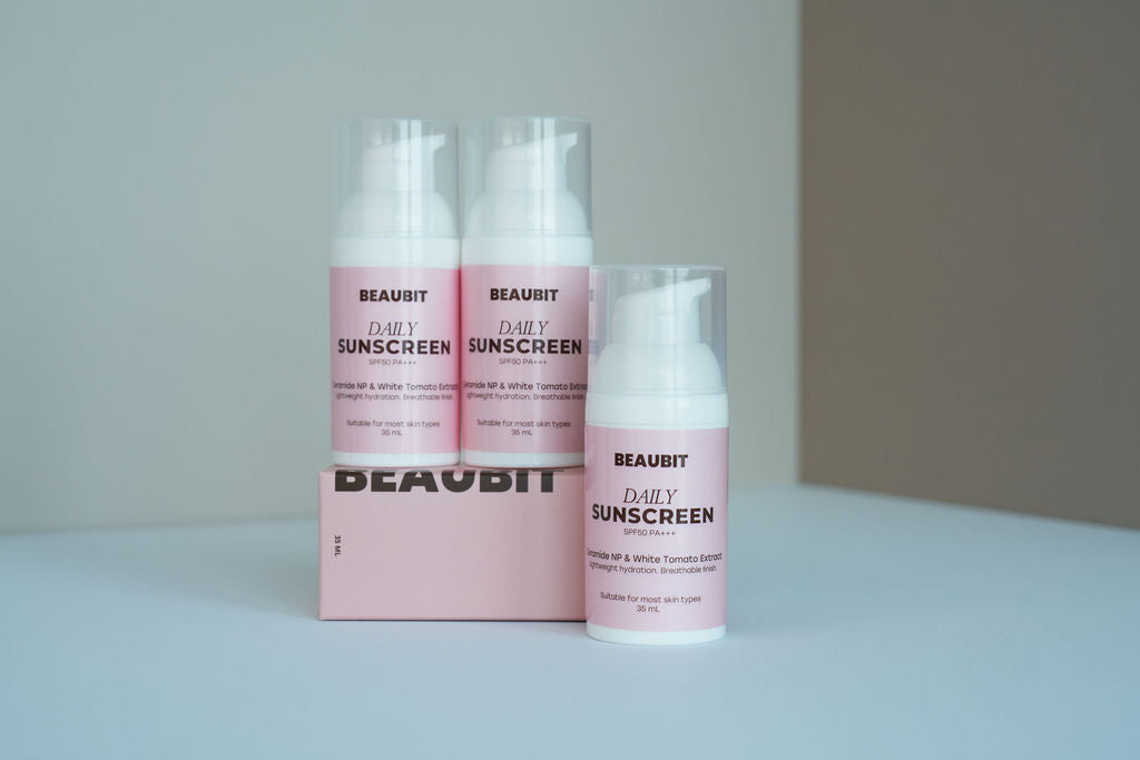 Three BEAUBIT Daily Sunscreen bottles and a matching pink box arranged on a light surface against a soft gray background.